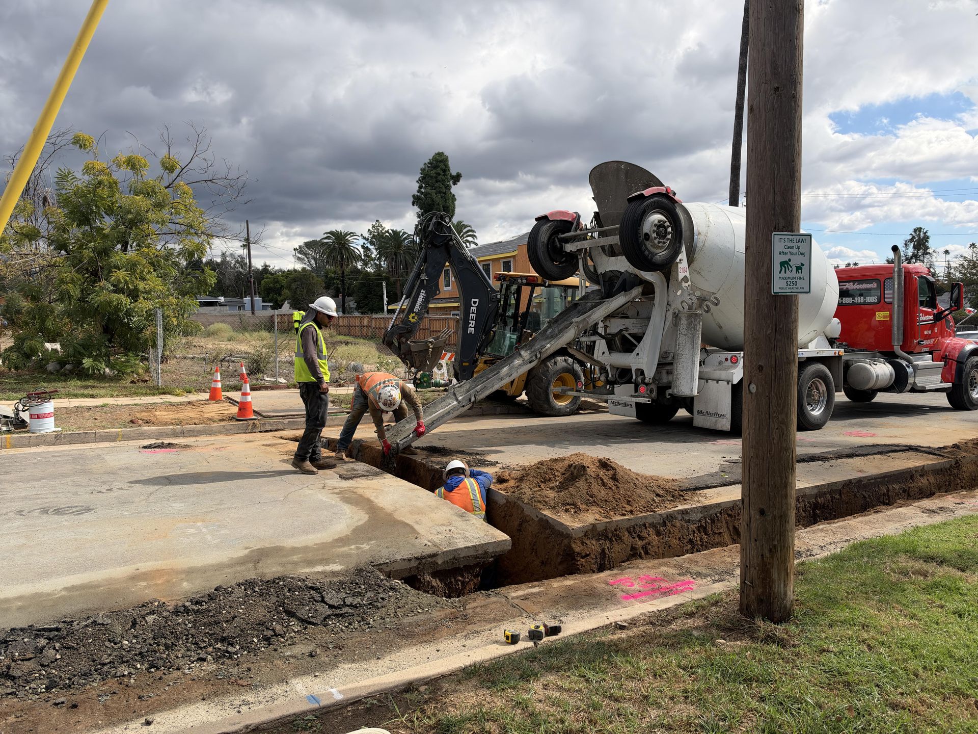 Full street view of a utility trench pour with mixer truck, backhoe, and crew